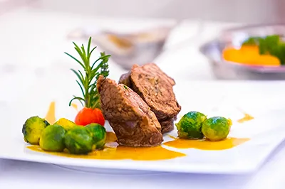 Kulinarische Produktfotografie: Gourmet-Speise auf Teller. Perfekte Präsentation für Menükarten und Restaurant-Websites. Fein angerichtetes Gericht auf weißem Teller mit Fleisch, Rosenkohl, Soße, Kirschtomate und Rosmarinzweig. Exzellente Food-Fotografie für die Gastronomie.